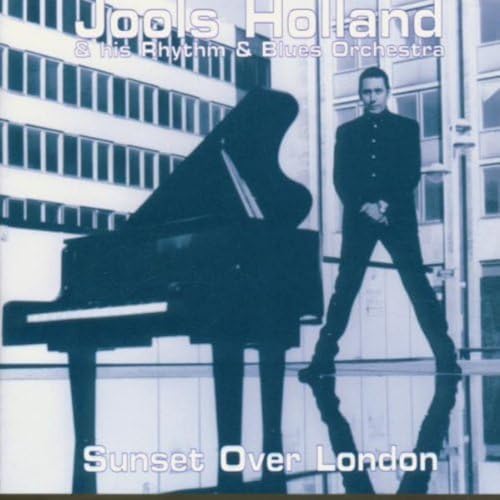 Jools Holland standing next to a grand piano against a blue-toned city building background.