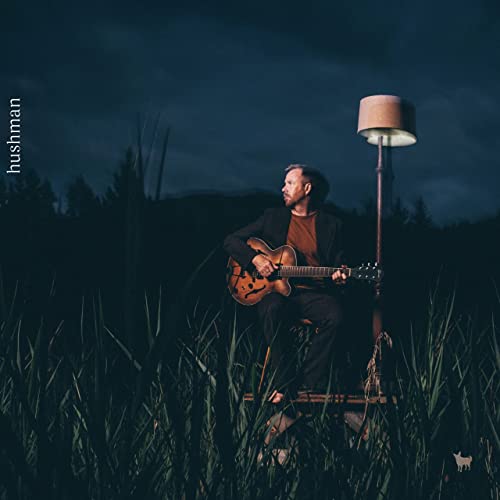 Man with guitar sits outdoors under a lamp, dark background.