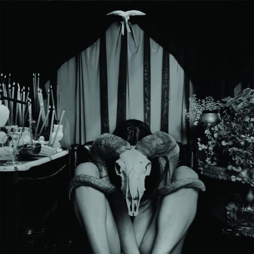 Person with ram skull mask, candles, and floral arrangements in a dark setting.