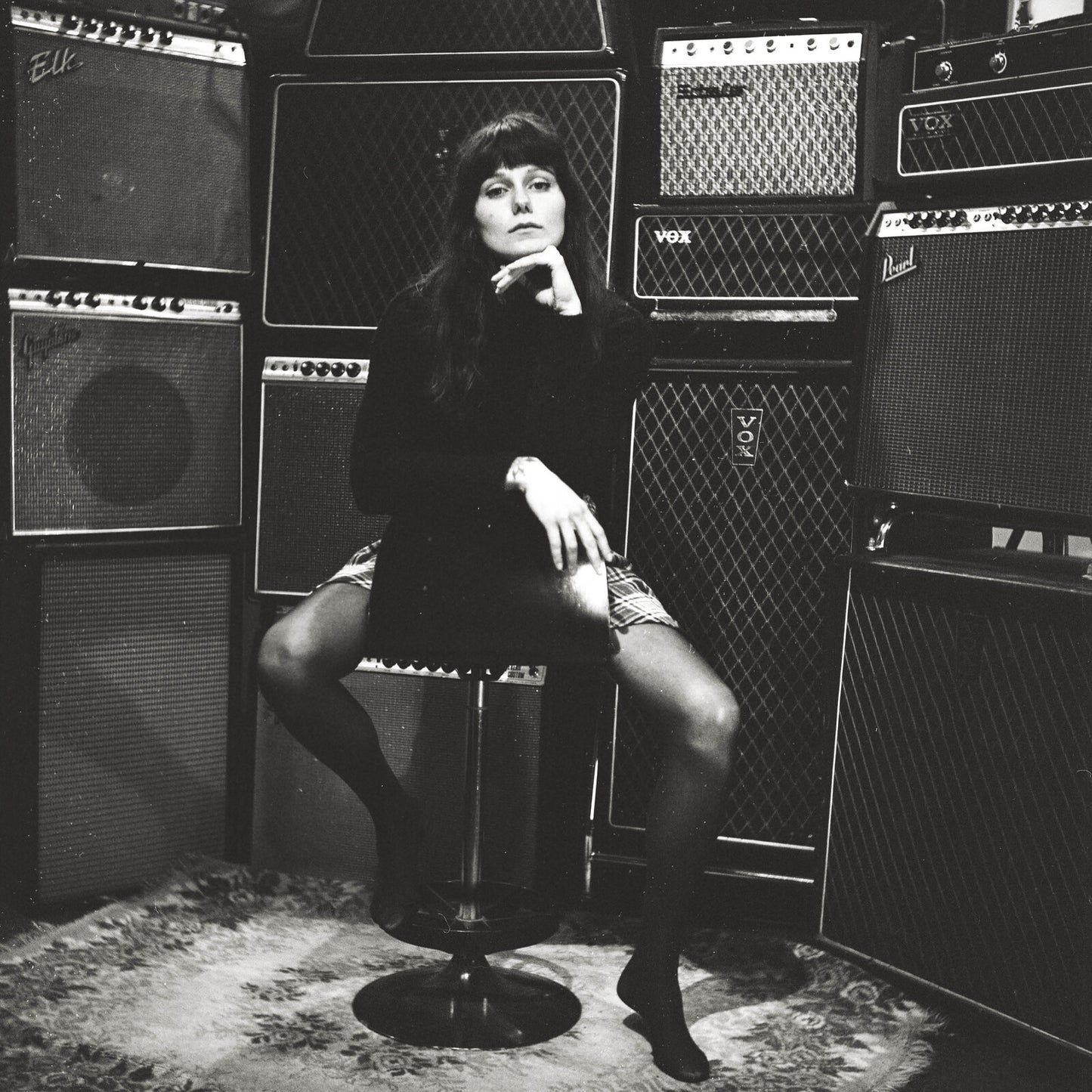 Black and white portrait of a woman seated amongst guitar amplifiers.