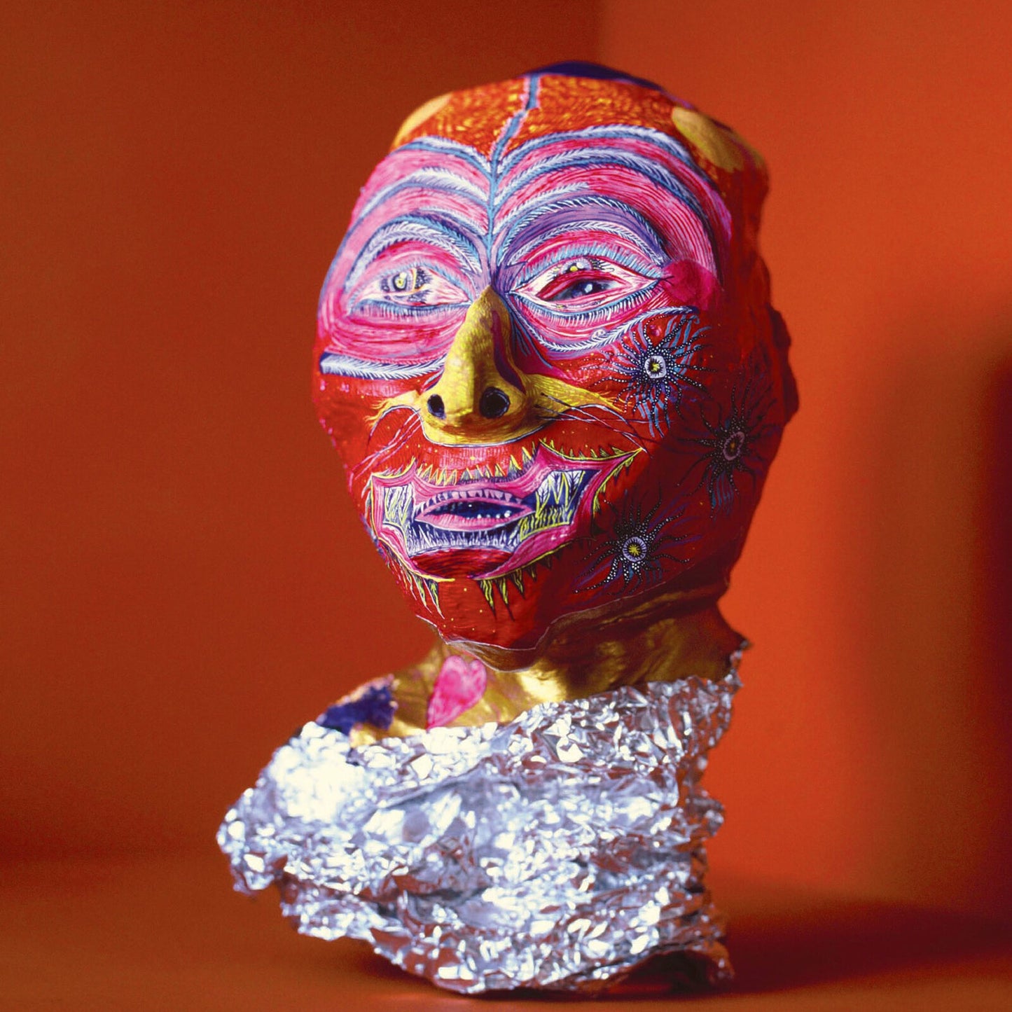 Painted human face sculpture with foil collar on a red background.