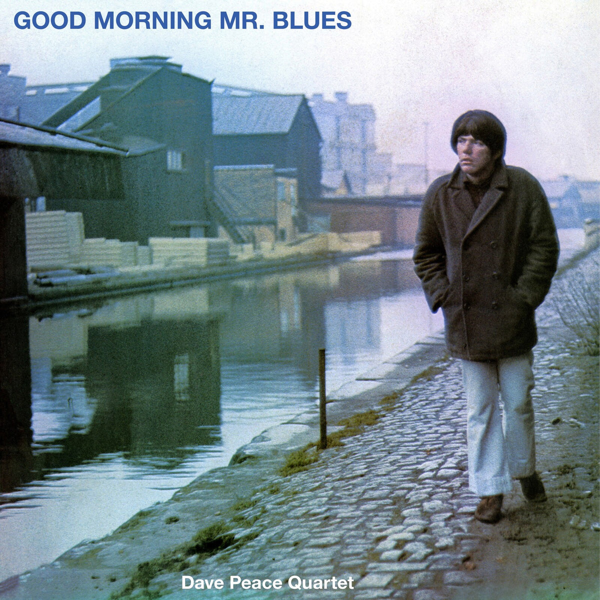 Album cover: man in coat by a canal, industrial buildings backdrop.