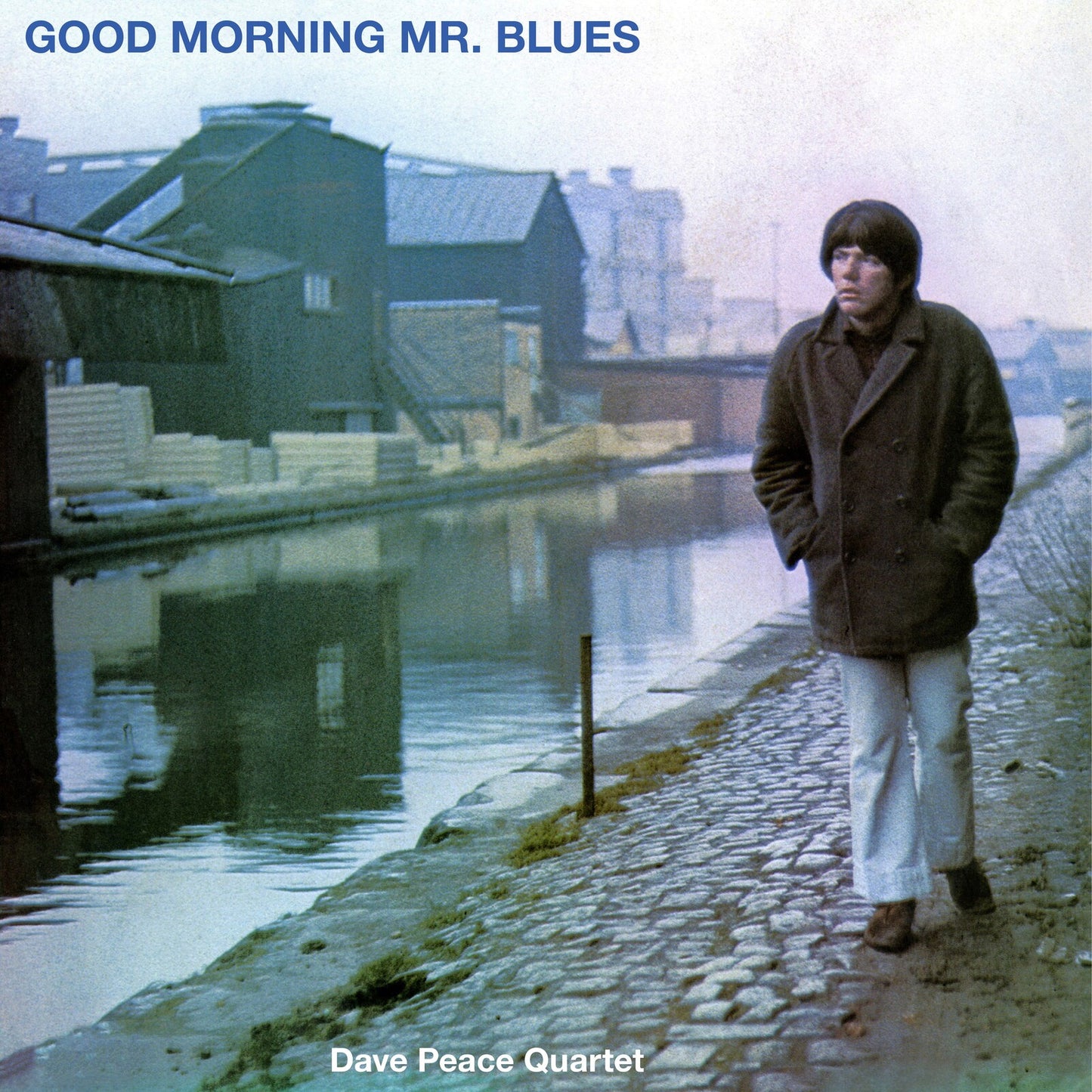 Album cover: man in coat by waterway, industrial buildings backdrop.