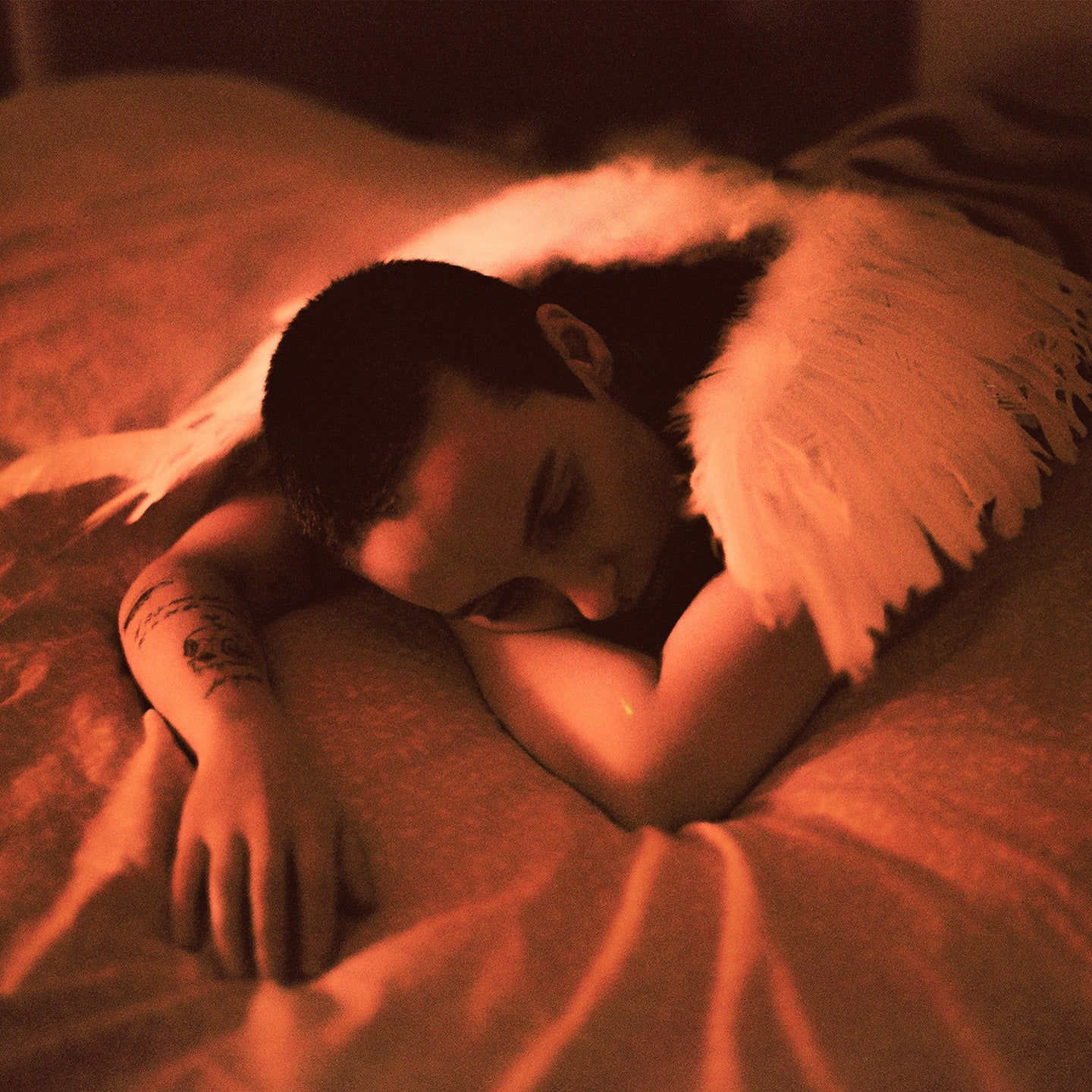 Person with tattoos lying on a bed, covered with a white blanket.