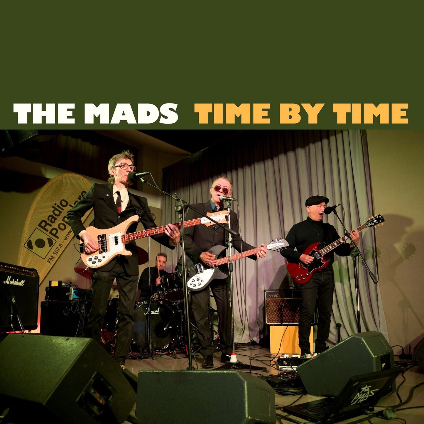 Band performing on stage with instruments, titled "The Mads - Time By Time.