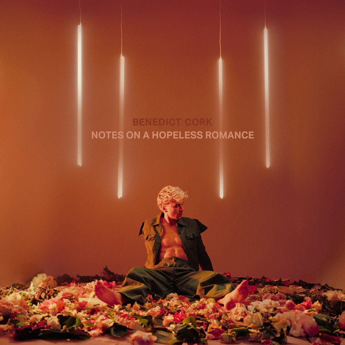 Man with blonde hair sits amid rose petals, lit by hanging lights.