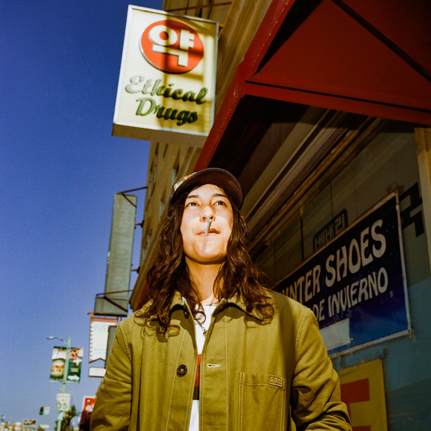Long-haired person in a jacket and cap stands in front of shops.