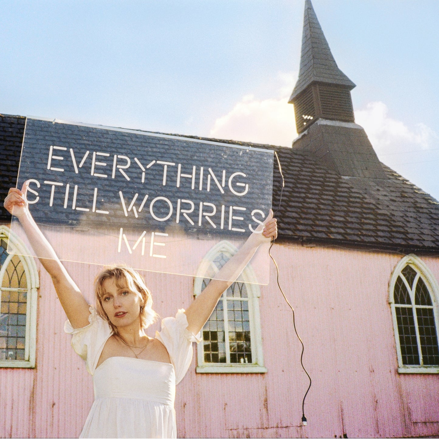 Woman in white dress holds sign reading “Everything Still Worries Me.”