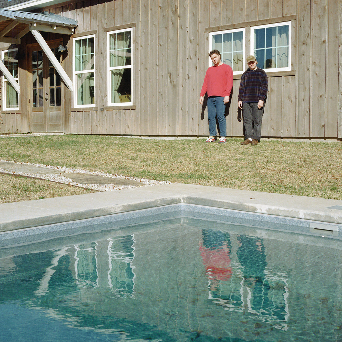Two men stand on grass near a wooden building and pool.