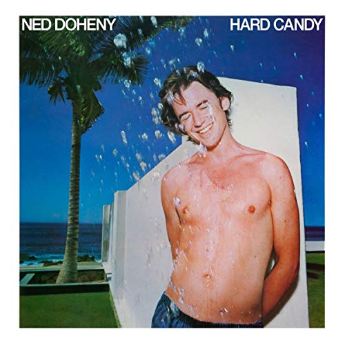 A shirtless man stands under splashing water near a white wall and palm tree.