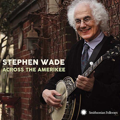 Stephen Wade with a banjo, smiling, against a brick wall background.