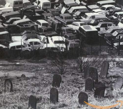 Black and white photo of a car junkyard with gravestones in foreground.