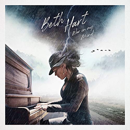 Beth Hart wearing a hat plays a piano under a stormy sky with lightning.
