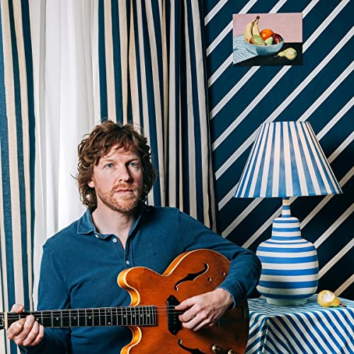 Man with guitar poses in a room with patterned wallpaper.