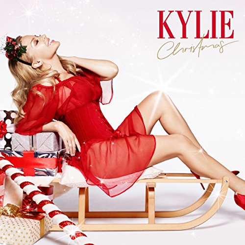 Kylie Minogue in red dress on a wooden sleigh, Christmas gifts nearby.
