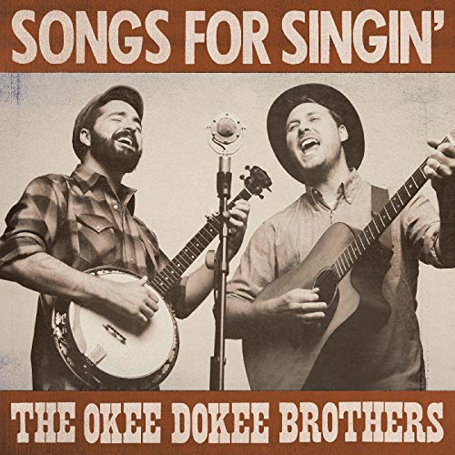 The Okee Dokee Brothers singing into a microphone while playing banjo and acoustic guitar.