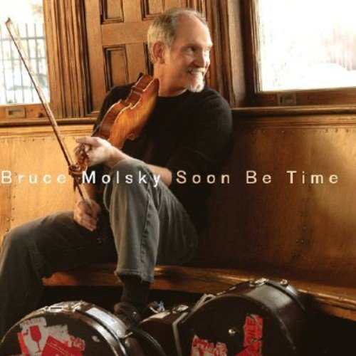 Bruce Molsky sits on a wooden bench holding a fiddle and bow.