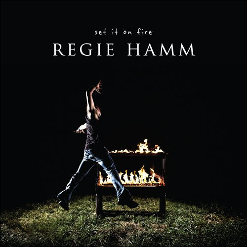 Album cover: person near burning metal box in dark field.