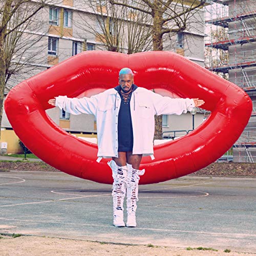 Man with blue hair and white boots standing before a giant red inflatable lip.