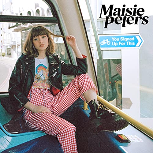 Maisie Peters wearing a leather jacket and red checkered pants sitting on a bus.
