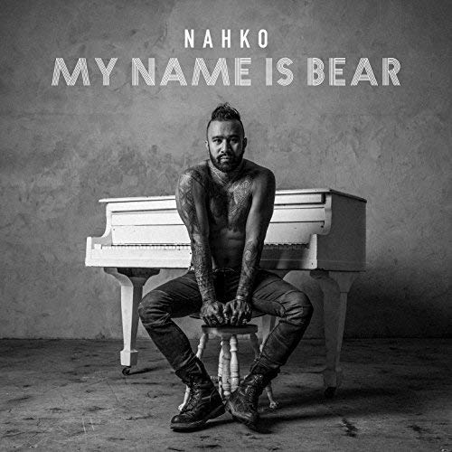 Nahko, shirtless with tattoos, sits before a white piano.