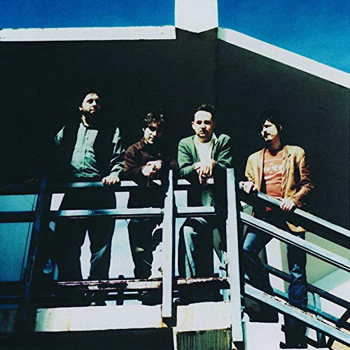 Four men stand on a rooftop railing against a blue sky.