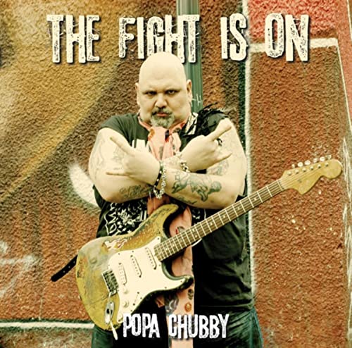 Popa Chubby holding an electric guitar in front of a textured wall.