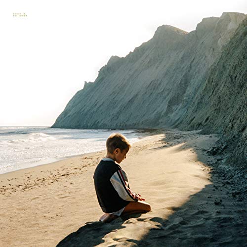 Boy sitting on a beach, facing a large cliff and ocean.
