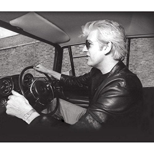 Black and white photo of Nick Lowe driving a vehicle, wearing sunglasses.