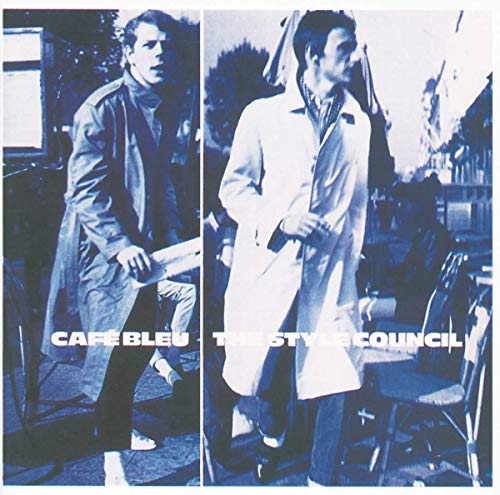 Two men in trench coats stand in a blue-toned, split-panel outdoor cafe scene.