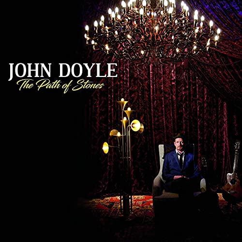 John Doyle sits in a chair before dark red curtains beneath a large chandelier.