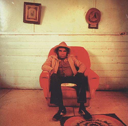 Man in a hat sits in a red armchair against a pale wall.