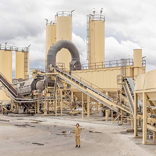 Woman stands before a large, industrial factory under a cloudy sky.