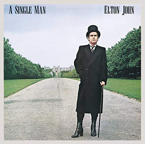Elton John standing in a long driveway wearing a top hat and black coat.