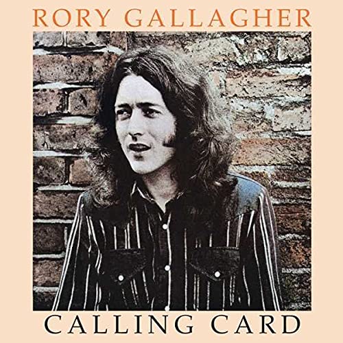 Rory Gallagher with long hair wearing a striped shirt in front of a brick wall.