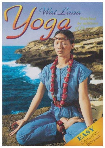 Woman in blue outfit and red lei meditates on rocky coastline with ocean.
