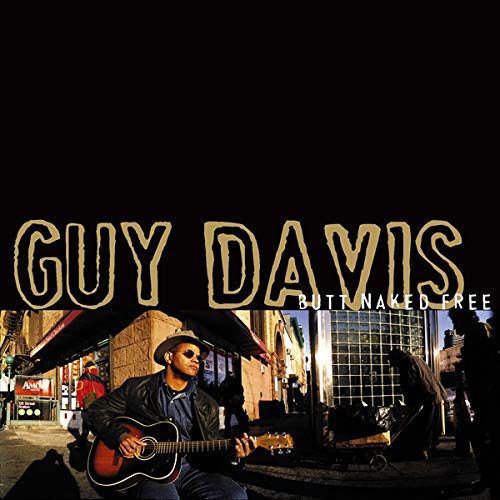 Guy Davis playing an acoustic guitar on a city street at sunset.