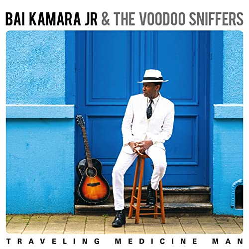 Man in white suit with guitar, seated before a blue door.
