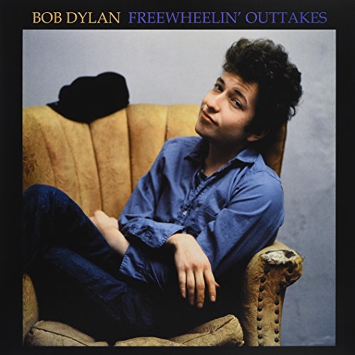 Bob Dylan sits cross-legged in front of a yellow armchair, wearing blue.
