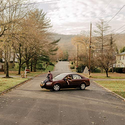 Two people sit on the hood of a maroon car on a street.