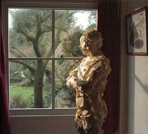 Figure covered in paper stands near a window overlooking a garden.
