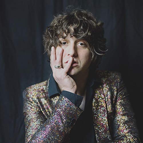 Curly-haired man in patterned jacket rests chin on hand.