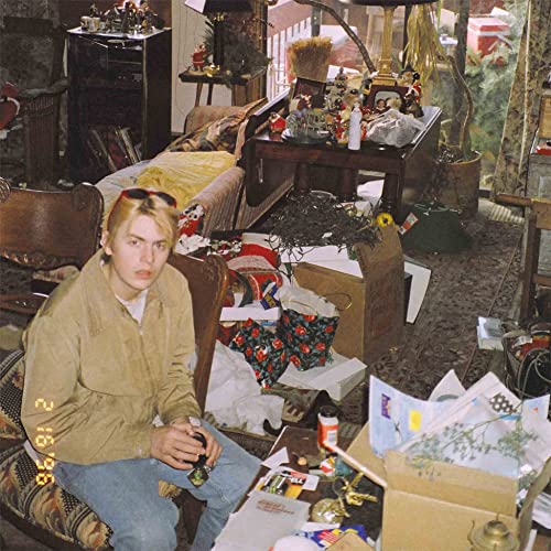 Woman in a tan jacket sits in a cluttered room with gifts.