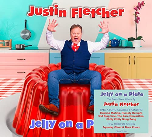 Justin Fletcher sits on a large red jelly, smiling with hands raised.
