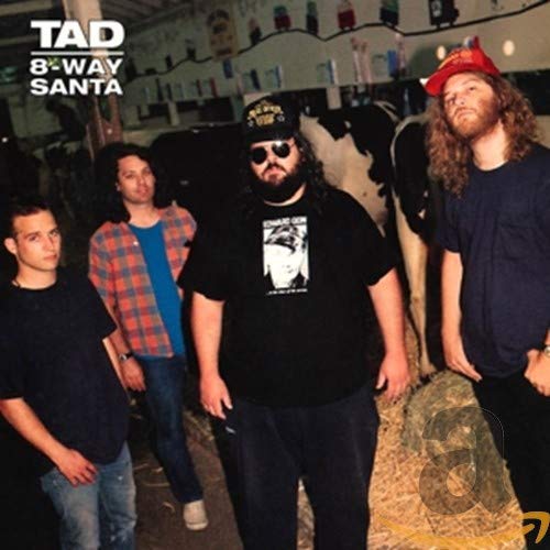 Four men stand in a barn with cows and hay bales.