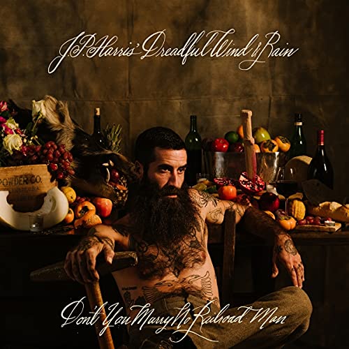Bearded man with tattoos sits amongst fruit, flowers, and bottles.