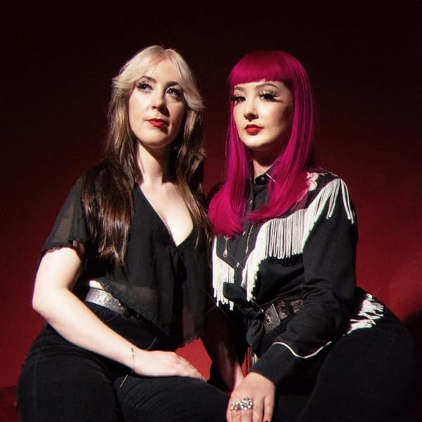 Two women with brightly colored hair pose against a dark red background.