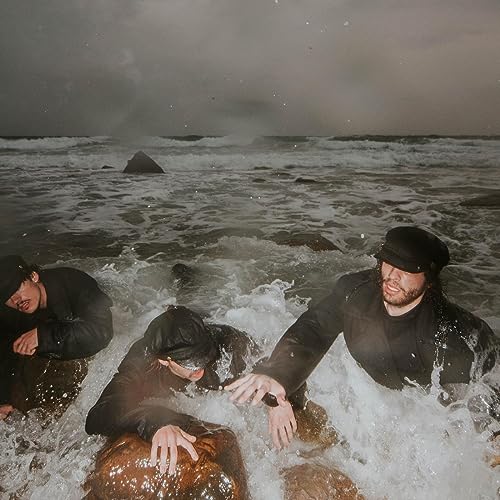 Three figures in dark clothing wade in stormy ocean waves.