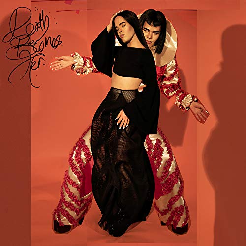 Two people in dramatic outfits pose against a reddish-orange background.