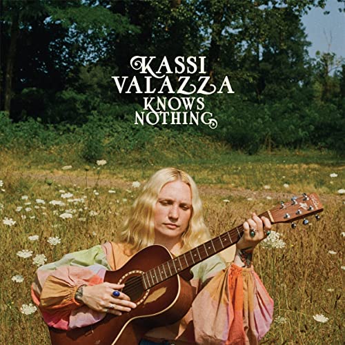 Kassi Valazza sits in a field, playing an acoustic guitar.
